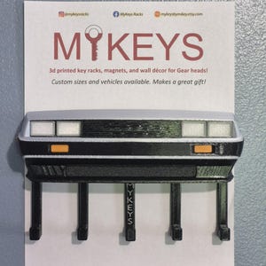 May include: A 3D printed key rack shaped like the front of a silver and black car with rectangular headlights and orange turn signals. The rack features five black hooks, with the center hook embossed with the text MYKEYS.