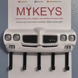 May include: A white 3D-printed key rack in the shape of a car's front, with black key hooks. It has headlights, a grill, and the text "GTO". The top displays "MYKEYS" and social media details for the seller.