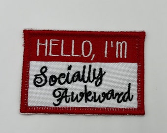 Peut inclure: Étiquette de nom en tissu rouge et blanc avec le texte "HELLO, I'M Socially Awkward" en broderie noire.