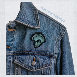 Puede incluir: Una chaqueta vaquera azul con un parche punk rock negro y azul claro con una calavera con un mohawk. El parche dice "Fancy Pants Patches" en una fuente azul claro.