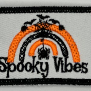 Può includere: Una toppa ricamata arancione e nera con una ragnatela e il testo "Spooky Vibes".