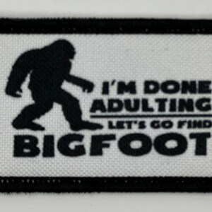May include: A rectangular embroidered patch with a white background and a black border. It features a black silhouette of Bigfoot and the text "I'M DONE ADULTING LET'S GO FIND BIGFOOT" in bold black lettering, arranged in three distinct lines.