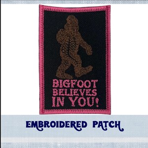 May include: Embroidered patch with a pink border. The patch features a brown silhouette of Bigfoot walking with the text "Bigfoot Believes In You!" in pink.