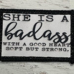 May include: Black and white embroidered patch with the text "She is a badass with a good heart, soft but strong."