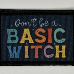 Op de afbeelding: Een rechthoekige stoffen patch met een donkerblauwe achtergrond en een zwarte gestikte rand. De tekst "Don't be a BASIC WITCH" staat in het midden gedrukt in een meerkleurig, verweerd lettertype in tinten blauwgroen, geel, oranje, rood en paars.