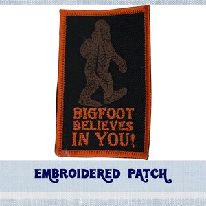 May include: Embroidered patch featuring a brown Bigfoot figure walking on a black background. The text "BIGFOOT BELIEVES IN YOU!" is in orange. The patch has an orange border.