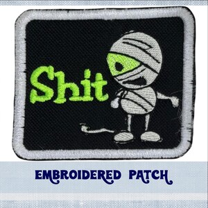 May include: Embroidered patch with a black background and white border. The patch features a green cartoon mummy with the word "Shit" in green letters.
