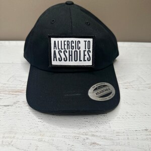 May include: Black adjustable baseball cap featuring the text "ALLERGIC TO ASSHOLES" on a white patch. The cap has a curved peak and a small button on top. The adjustable strap is visible on the back.