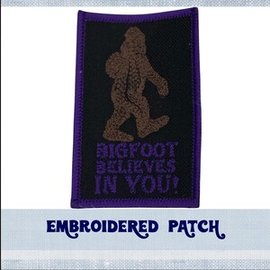 May include: Embroidered patch with a purple border. The patch features a brown Bigfoot walking with the text "BIGFOOT BELIEVES IN YOU!" in purple.