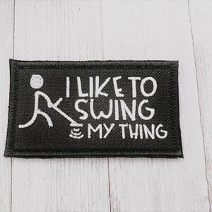 May include: Black rectangular patch with white embroidered text and a stick figure. The text reads "I LIKE TO SWING MY THING". The stick figure is swinging a golf club. The patch has a black border.