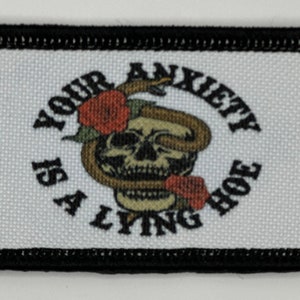 May include: Embroidered patch with a black border and a white background. The patch features a skull with a snake wrapped around it, and two red roses. The text "YOUR ANXIETY IS A LYING HOE" is arched around the skull.
