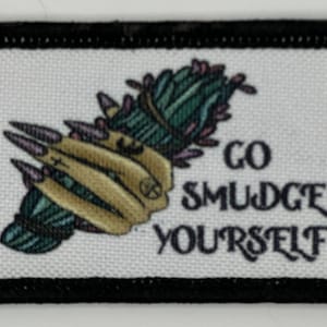 Puede incluir: Un parche blanco con costuras negras alrededor de los bordes. El parche presenta una ilustración verde, morada y amarilla de un palo de incienso con el texto "GO SMUDGE YOURSELF" en negro.
