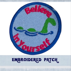 Puede incluir: Parche bordado con borde azul y fondo azul claro. El parche presenta un monstruo del Lago Ness verde con el texto "Believe in Yourself" en letras rosas.