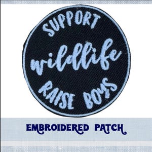 May include: A circular, black embroidered patch featuring the light blue text "SUPPORT wildlife RAISE BOYS". The words "EMBROIDERED PATCH" are written in dark blue script below the patch.