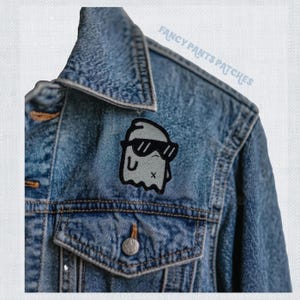 Könnte beinhalten: Ein gestickter Patch in Schwarzweiß, der ein Gespenst mit Sonnenbrille auf einer blauen Jeansjacke zeigt. Der Patch trägt die Aufschrift "Fancy Pants Patches" in weißer Schrift.