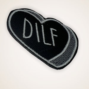 May include: A black heart-shaped patch with the word "DILF" embroidered in white thread. The patch has a gray border and is set against a white background.