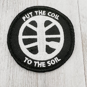 Könnte beinhalten: Schwarz-weiß gestickter Patch mit dem Text "Put the coil to the soil" und einem stilisierten Kreisdesign.