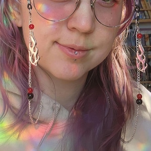 Puede incluir: Cadenas de gafas plateadas con colgantes de llama, cuentas rojas y negras. Las cadenas están unidas a gafas redondas con montura plateada. La persona de la imagen tiene el pelo morado y un piercing en el labio. El fondo está borroso.