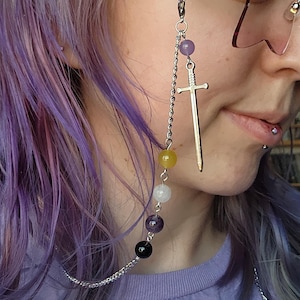 May include: A silver-toned metal eyeglass chain featuring a series of round beads in shades of purple, yellow, white, and black. The chain includes a silver-colored sword charm and attaches to the glasses with a lavender-colored silicone loop.