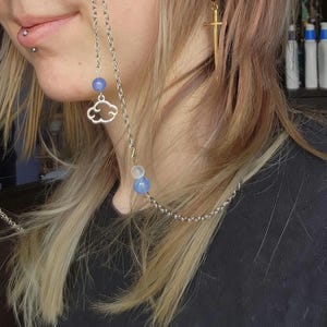 May include: Silver chain glasses holder featuring a cloud charm and blue beads. A gold sword earring with a red bead is also visible. The jewellery is worn by a person with blonde hair and a black top.