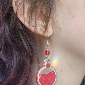 May include: A clear, round earring featuring a red liquid design, red beads, and a silver hook. The earring is shown near an ear, with purple hair in the background.