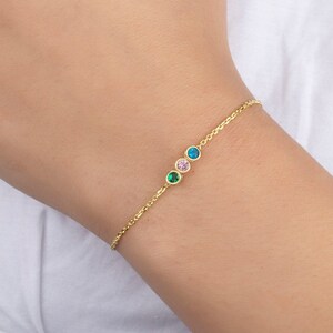 Op de afbeelding: Een gouden kettingarmband met drie kleine ronde edelstenen in groen, roze en blauw.