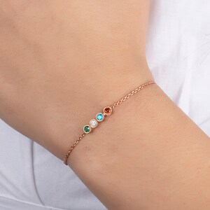 Op de afbeelding: Een delicate gouden kettingarmband met vijf kleine ronde edelstenen in verschillende kleuren: groen, blauw, wit, turquoise en rood.