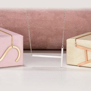May include: Three initial necklaces: a gold 'S', a silver 'T', and a rose gold 'A'. Each letter is large and attached to a fine chain. The necklaces are presented on neutral boxes, set against a pink backdrop.