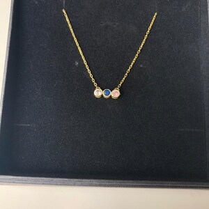 Op de afbeelding: Een gouden ketting met drie kleine ronde stenen. De stenen zijn wit, roze en blauw.