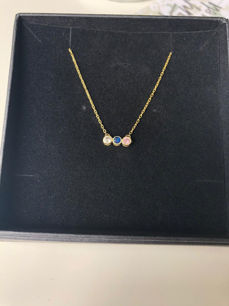 Op de afbeelding: Een gouden ketting met drie kleine ronde stenen. De stenen zijn wit, roze en blauw.