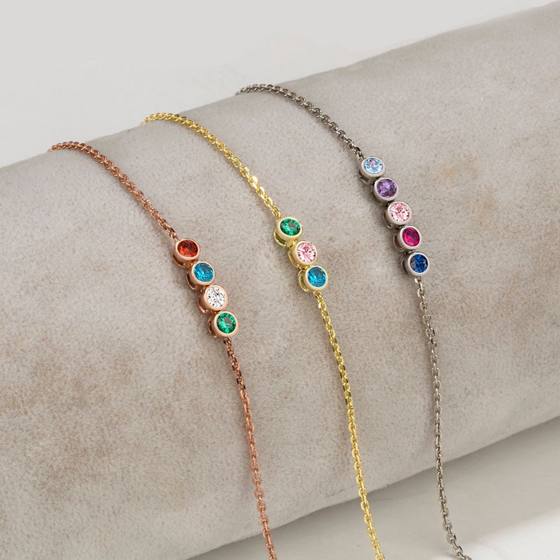 Op de afbeelding: Vier delicate armbanden met een kettingontwerp en kleine gekleurde edelstenen. Elke armband heeft een andere kleurencombinatie van edelstenen, waaronder blauw, groen, roze en rood.