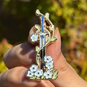 May include: A gold-toned metal enamel pin featuring a silver sword with a blue handle grip. The sword is entwined with light green vines and surrounded by clusters of small, light blue five-petaled flowers with gold centers.