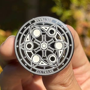 May include: A circular black and white enamel pin featuring a central geometric flower pattern surrounded by six eye-shaped motifs. The outer border contains small star symbols and the text ZOLTRAAK printed twice along the top and bottom edges.