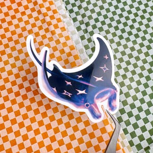 May include: A sticker featuring a stylized illustration of a manta ray. The manta ray is primarily dark blue with pink and white accents, and star-shaped details. The sticker is held by tweezers against a checkered orange and green background.
