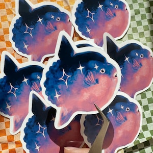 May include: A collection of stickers featuring a stylized ocean sunfish design. The stickers showcase a gradient of pink and blue hues, with sparkling star accents. The sunfish has a distinctive shape and is set against a white background.