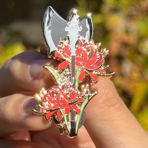 May include: A metal enamel pin featuring a silver double-bitted axe overlaid with two vibrant red spider lily flowers. The design includes gold-toned metal outlines and small star-shaped accents, creating a detailed botanical and weapon-themed accessory.