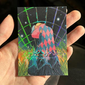 May include: A rectangular holographic print featuring a couple embracing under a starry night sky. The figures are depicted in a colorful, stylized manner, with one wearing a harlequin-patterned cloak. The print has a dark background with vibrant green and orange accents.