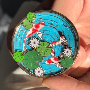 May include: A circular enamel pin featuring a vibrant blue glitter background representing water, with three koi fish in red, white, and black patterns swimming among green lily pads and small white water lily flowers with gold metal outlines.