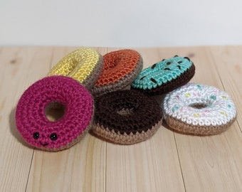 Puede incluir: Seis donuts de crochet en varios colores, incluyendo rosa, amarillo, naranja, marrón, blanco y azul. Los donuts tienen una cara sonriente y están dispuestos sobre una superficie de madera.