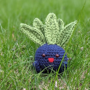 May include: A blue and green crocheted plush toy with a smiling face. The toy is sitting in a bed of green grass.