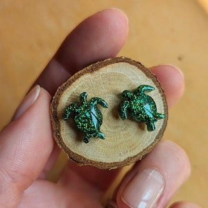 May include: A pair of green glitter turtle earrings on a wooden surface.