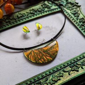 May include: A set of jewellery featuring a necklace and earrings with a crescent-shaped pendant and matching earrings. The pendant and earrings are made of clear resin with a pressed green and yellow leaf design.