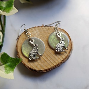 May include: A pair of silver earrings with a white and green iridescent moon and a silver hare silhouette.