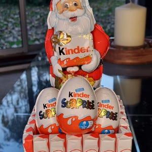 May include: A Christmas-themed gift arrangement with a Santa Claus figure holding a Kinder chocolate bar, Kinder Surprise eggs, and chocolate bars. The arrangement is decorated with a red bow.