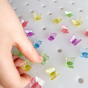 May include: A collection of small, colorful butterfly-shaped embellishments. The butterflies are translucent and come in various colors, including red, purple, blue, green, and yellow. The image shows a hand interacting with the butterflies, which are scattered on a white surface.