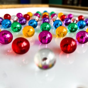 May include: A collection of colorful, shiny glass marbles in various colors, including red, pink, green, blue, yellow, and silver. The marbles are arranged on a reflective white surface, creating a vibrant and playful display.