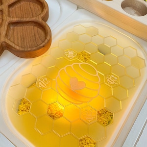 May include: A yellow honeycomb-shaped tray with a white beehive and bees etched into the surface. The tray is filled with yellow crinkle paper and is illuminated from below.
