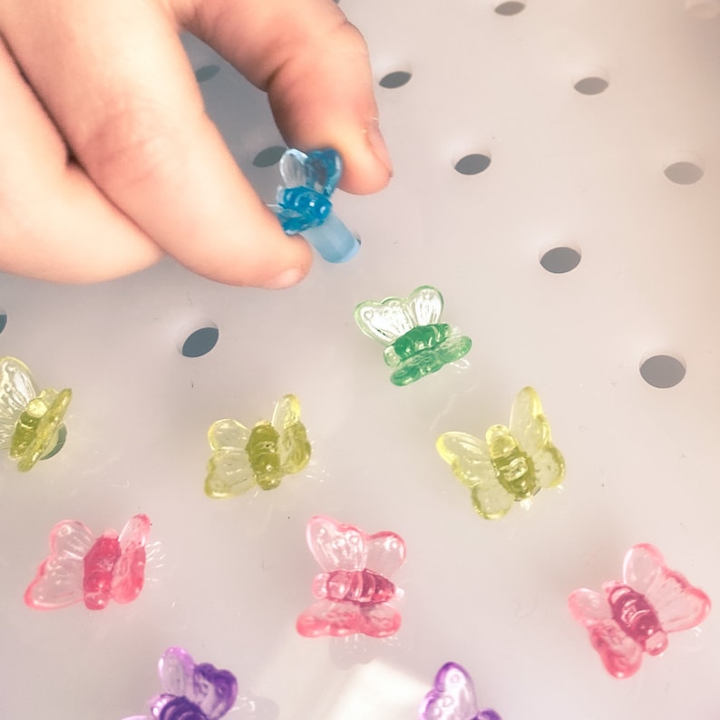 May include: A hand holding a translucent blue butterfly-shaped bead. Several other colorful butterfly beads in shades of green, yellow, pink, and purple are scattered on a white surface. The beads have a glossy finish.