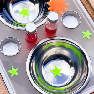 May include: A sensory play setup featuring two stainless steel bowls with neon green star accents, small jars with red liquid, and white powder in small metal containers. An orange sun-shaped object and neon green stars are scattered around.