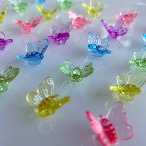 May include: A collection of small, translucent butterfly-shaped embellishments in various colors, including pink, blue, green, yellow, and purple. The butterflies are scattered across a reflective surface, creating a colorful and whimsical display.